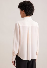Woman with curly hair wearing a long-sleeve, light beige blouse and black pants, shown from the back against a plain background.