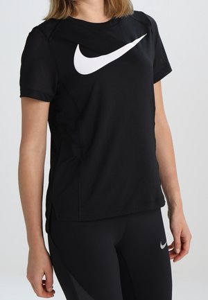 Femme portant un t-shirt Nike noir avec un grand logo blanc et un legging Nike noir assorti, debout devant un fond clair uni.