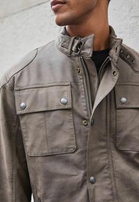 Chaqueta de cuero marrón con dos bolsillos frontales, herrajes plateados, botones a presión y un cuello alto. Cierre de cremallera que se extiende por el centro.