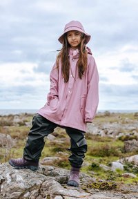 Imperméable rose et étanche avec poches et fermetures à pression, associé à un pantalon noir imperméable et des chaussures texturées navy, se tient sur des rochers en extérieur.
