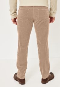 Beige Cordhose mit geradem Schnitt, zwei Gesäßtaschen und glatter Textur. Kombiniert mit dunkelbraunen Schuhen.