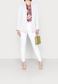 Blazer gris clair porté sur un haut à motif floral, assorti à un jean skinny blanc et à des sandales à talons beiges. Tient un petit sac à main métallisé doré.
