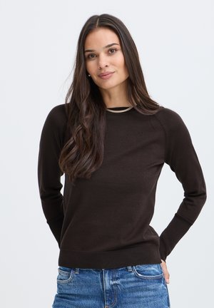 Jeune femme aux longs cheveux foncés portant un pull noir à manches longues et un jean bleu, debout les mains dans le dos sur fond blanc.
