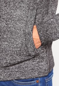 Grey knitted sweater with a textured pattern, featuring ribbed cuffs and a side pocket, paired with blue denim jeans.