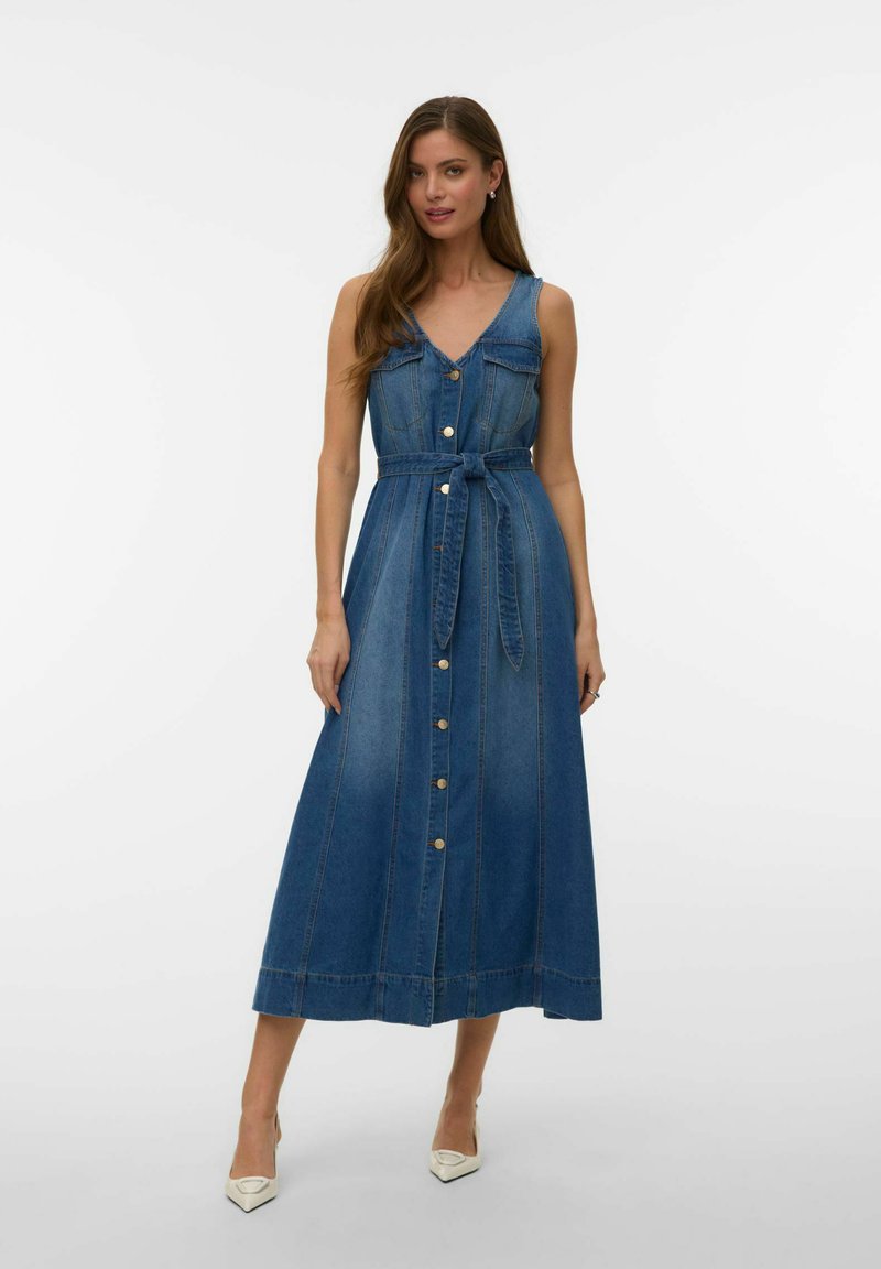 Femme portant une robe midi en denim bleu sans manches avec des boutons à l'avant et une ceinture à la taille, debout en talons blancs sur un fond uni.