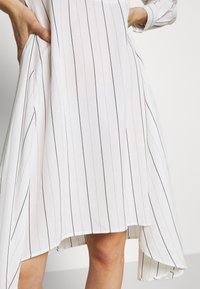 Abito bianco con strisce verticali nere, caratterizzato da un design fluido e un orlo asimmetrico. Realizzato in tessuto leggero.