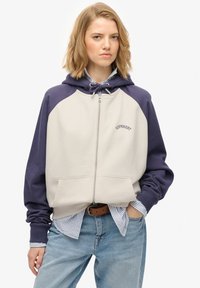 Felpa con zip in colorazione crema e blu navy, caratterizzata da una tasca a marsupio, cappuccio con coulisse e logo ricamato "SUPERDRY" sul petto.