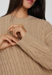 Beige Wollpullover im Cable-Knit-Stil mit rundem Halsausschnitt, gerippten Bündchen und einem strukturierten Muster. Hände passen den Ärmel an.