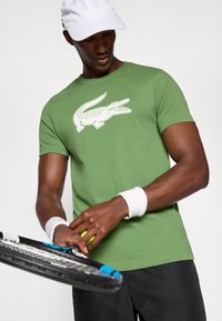 Zaļa kokvilnas T-kreklā ar baltu krokodila logo, īsām piedurknēm, apvienots ar melniem šortiem un plaukstas siksnām, tur rokās tenisa raketi.