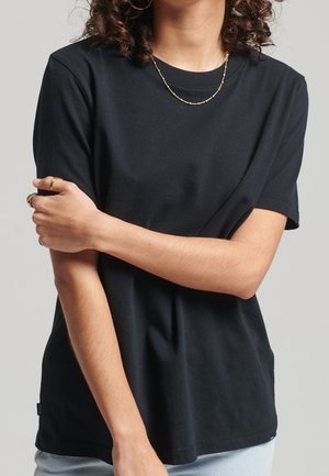 Person wearing a plain black short-sleeve shirt and gold necklace, with arms crossed over the torso against a plain light background.