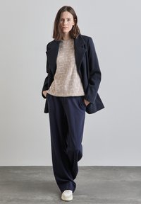 Marinblå kavaj över en beige texturerad tröja med geometriska mönster, matchad med lösa marinblå byxor och vita sneakers på en grå bakgrund.