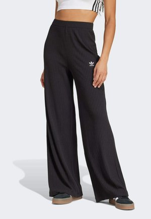 Femme portant un pantalon noir à larges jambes côtelé avec logo Adidas, associé à des baskets noires et un top court blanc.