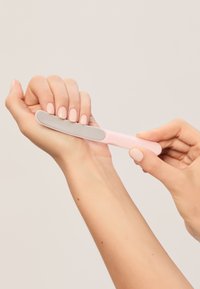 Vaaleanpunainen manikyyrityökalu, jossa on kaareva reuna; siinä on sileä metallinen hiontapinta. Käsi pitää sitä ja kauniisti hoidettuja kynsiä neutraalia taustaa vasten.