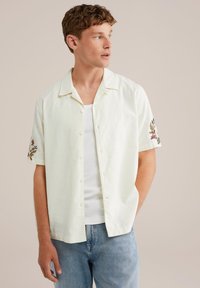 Camisa de manga corta de color crema claro con cuello, que presenta bordados florales en las mangas y cierre de botones, combinada con jeans azules claros.