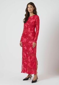 Rød kjole med lange ærmer, blomsterbroderi, transparent overflade og sideslidser. Bæres med sorte slingback-højhælede sko. Glat tekstur.