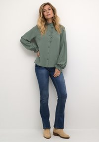 Camisa de manga larga y botones en verde apagado con cuello y puños con volantes, combinada con vaqueros azules y botines beige. Textura de tejido suave.