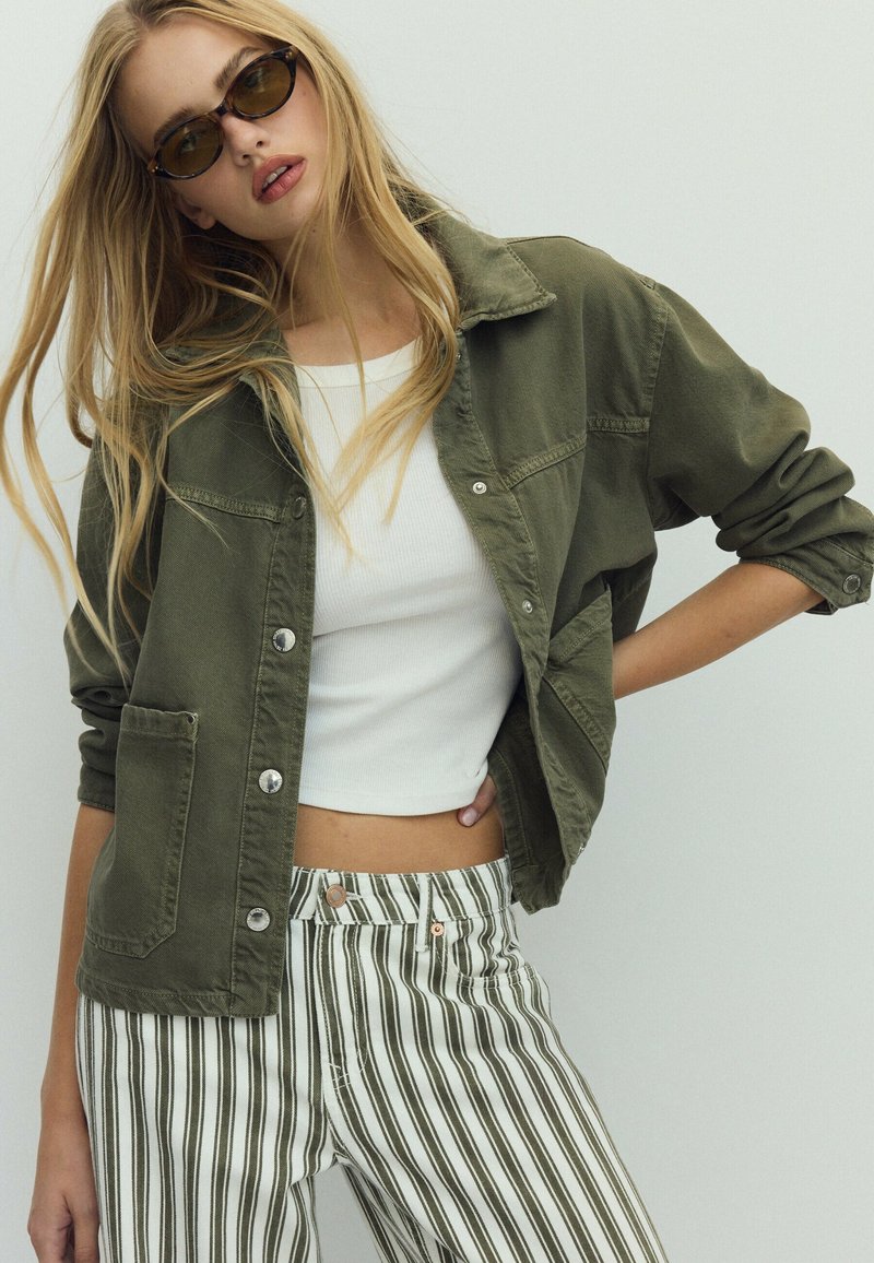 Femme portant des lunettes de soleil, une veste vert olive, un crop top blanc et un pantalon blanc à rayures noires verticales, posant avec la main sur la hanche.