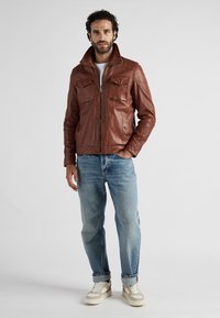 Brun läderjacka med dragkedja framtill, två bröstfickor och en strukturerad krage; bärs över en vit t-shirt och blå jeans.