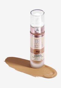 Botella de base de maquillaje de larga duración Revolution IRL Filter en el tono F12, presentada en un dispensador de bomba transparente; muestra de líquido beige cálido sobre la superficie.