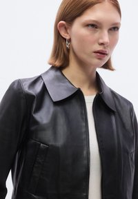 Μαύρο δερμάτινο μπουφάν με λεία υφή, που διαθέτει φαρδύ γιακά, μπροστινό φερμουάρ και πλαϊνή τσέπη. Απλά, σύγχρονα στοιχεία σχεδιασμού.