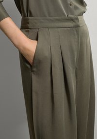 Main droite d'une femme reposant à l'intérieur d'une poche plissée de pantalons vert olive avec une taille large, tandis qu'une manche de chemise boutonnée est visible.