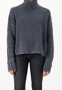 Pull à col roulé rayé dans des teintes bleu marine et gris, avec une coupe oversize, une texture côtelée et des manches allongées. Associé à un jean sombre.