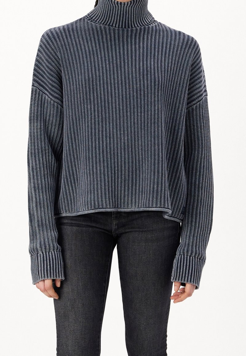 Pull à col roulé rayé dans des teintes bleu marine et gris, avec une coupe oversize, une texture côtelée et des manches allongées. Associé à un jean sombre.