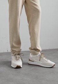 Adidași albi cu accente bej și șireturi texturate, având o talpă groasă și un design subțire, aerodinamic.