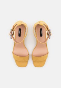 Keltaiset mokkanahkaiset korkokenkäsaappaat, joissa on leveä etuhihna, avonainen takaosa ja koristeellinen solki, jossa on monivärinen kuvio.