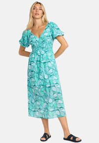 Une robe maxi fleurie turquoise avec des manches courtes bouffantes, un décolleté en V profond et une taille élastique, assortie à des sandales sombres.