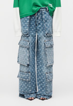 Blå cargobukser i vid benstil i denim med flere store lommer med klapper og et subtilt monogrammønster over det hele, båret med en grøn og hvid lagdelt top.