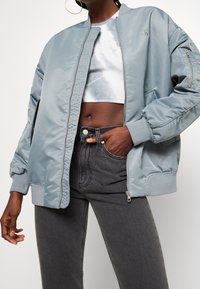 Giacca bomber blu chiaro con finitura satinata liscia, polsini a coste e tasca con zip. Indossata sopra un top corto metallizzato e jeans scuri.
