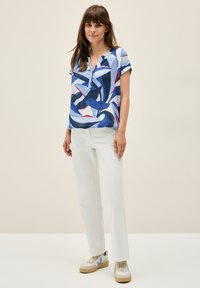 Camicia a manica corta con un motivo astratto blu, bianco e rosso; presenta un colletto a V. Abbinata a pantaloni bianchi e sneaker beige.