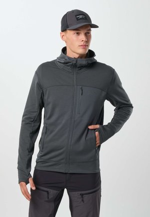Jeune homme portant une veste zippée gris foncé avec capuche et une casquette de baseball assortie, debout avec une main dans la poche de la veste.