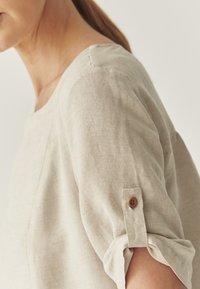 Blouse en lin beige avec un col rond, des manches roulées et un accent de bouton marron sur le poignet. Texture lisse avec un éclat minime.