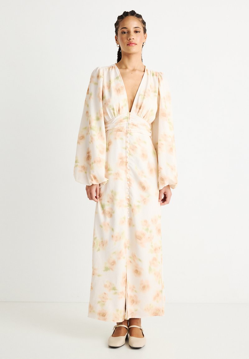 Woman with braids wearing a long cream dress with pastel floral print, deep V-neckline, button details, and puffed long sleeves, standing.