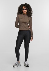 Braunes Langarmoberteil mit Reißverschluss am Hals, kombiniert mit schwarzen Leggings und grauen Turnschuhen. Das Outfit bietet ein figurnahes Design und eine glatte Textur.