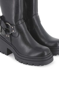 Botas de ante negras hasta el tobillo con un tacón grueso, textura grabada, cierre de cremallera y acentos de hardware plateado en la correa del tobillo.