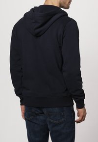 Hombre vistiendo una sudadera negra sencilla y jeans azul oscuro, visto desde atrás contra un fondo claro y liso.