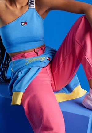 Jasnoniebieski top na jedno ramię typu crop z logo, w zestawieniu z różowymi dresami. Niebieska bluza z żółtymi akcentami jest zarzucona na biodra.