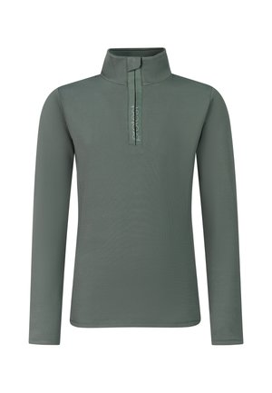 Junge Frau steht und trägt eine schwarze Mütze, ein grünes langärmeliges Zip-up-Top, schwarze Hose und schwarze Schuhe vor einfachem Hintergrund.