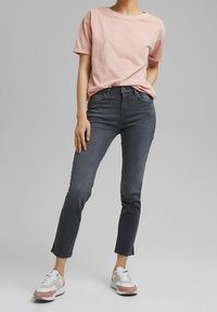 Persona que lleva una camiseta de manga corta rosa claro, jeans ajustados gris oscuro y zapatillas blancas con acentos rosas y beige, de pie de manera informal.