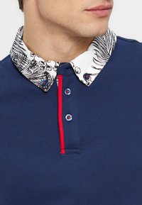 Άνδρας που φοράει μπλε μακρυμάνικη πόλο με λευκό γιακά με λευκό φλοράλ μοτίβο, τρία μπλε κουμπιά με κόκκινη ραφή και κόκκινη κάθετη ρίγα κατά μήκος του κουμπότρυπας.