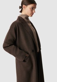 Bruine wollen jas met een losse pasvorm, geknipte revers en zijzakken. Draagt over een beige geribde trui met een gladde textuur en diepe kleur.