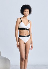 Γυναίκα στέκεται με λευκό σετ σουτιέν και εσώρουχο Calvin Klein με μαύρα λουριά και ζώνη μέσης, στο φόντο απλού ανοιχτού γκρι.