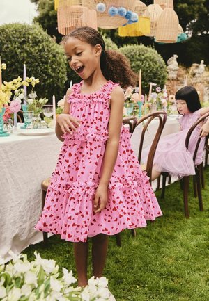 Fată într-o rochie roz cu model de inimioare stă lângă o masă decorată în aer liber, zâmbitoare, iar aproape este o altă fată așezată, îmbrăcată într-o rochie lavandă.