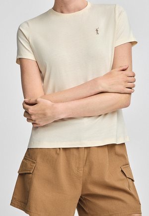 Osoba nosząca kremowy t-shirt z krótkim rękawem z małym logo na piersi i brązowe szorty cargo, ręce skrzyżowane na torsie.