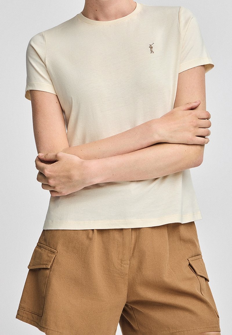 Personne portant un t-shirt crème à manches courtes avec un petit logo sur la poitrine et un short cargo marron, bras croisés sur le torse.