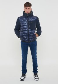 Marineblauwe gewatteerde jas met een hoge kraag, ritssluiting en borstzak; gecombineerd met blauwe jeans en zwart/witte sneakers.