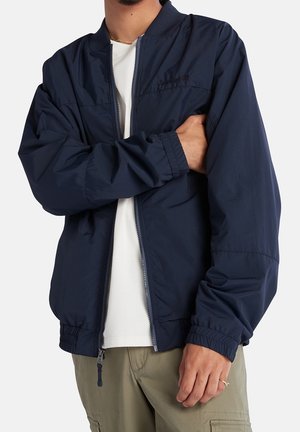 Homme portant une veste zippée bleu marine par-dessus une chemise blanche et un pantalon kaki, les bras croisés, une main tenant le bras opposé.